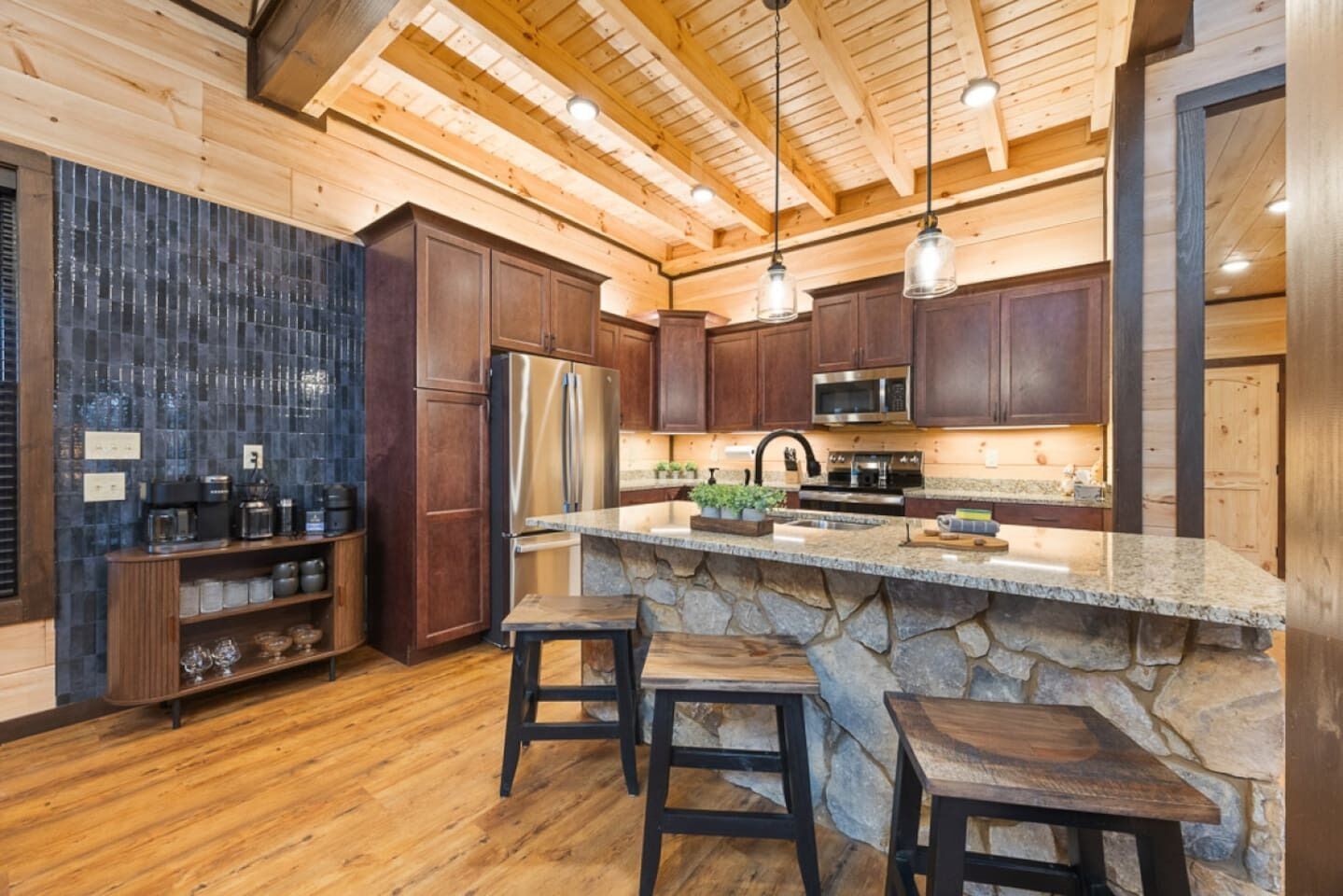 Une cuisine moderne avec des meubles de couleur sombre en bois, un grand îlot central doté de tabourets, un mur d’accent en pierre et un plafond en bois.