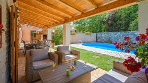 A covered outdoor patio with wicker furniture, a pool, and a stone wall.