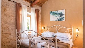 A bedroom with a stone wall, a bed with white metal headboard, and a painting of leaves on the wall.
