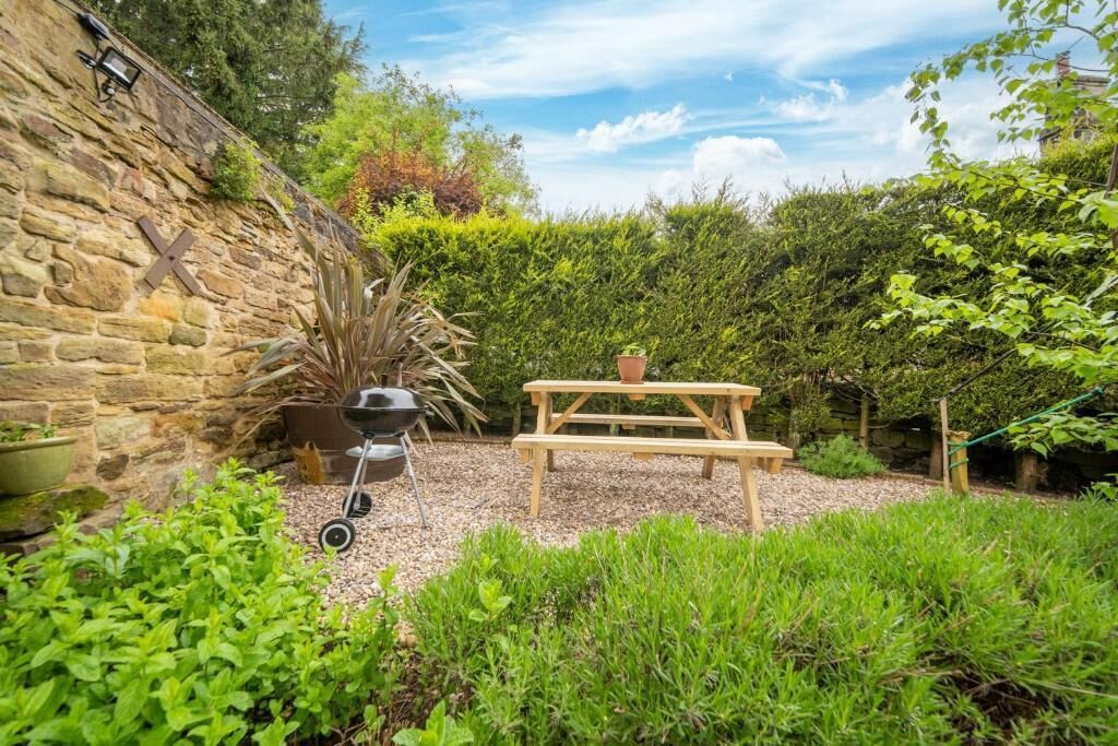 A stone wall, a barbecue grill, and a wooden picnic table in a garden with dense greenery.