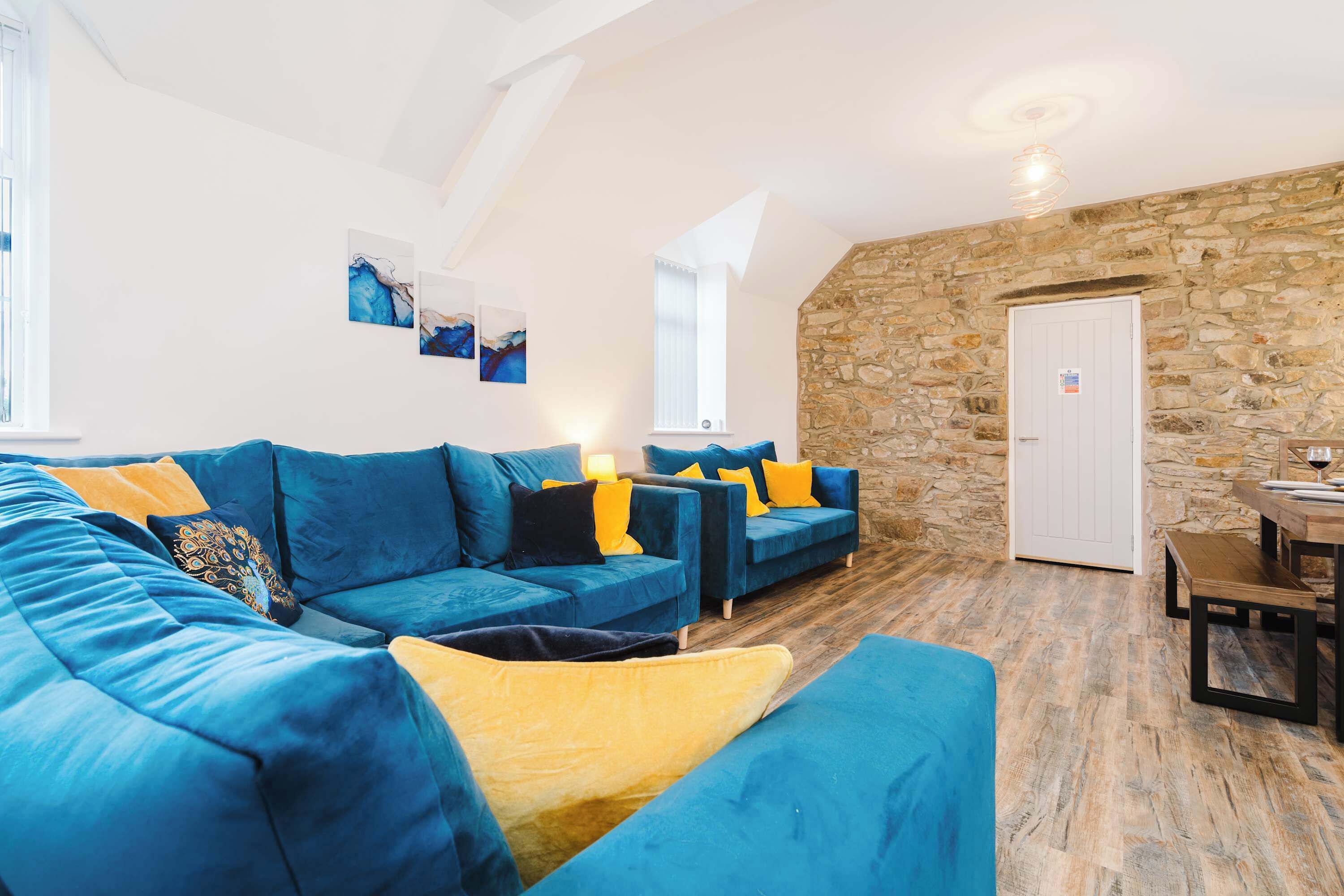 A modern living room with a blue sofa set, a stone wall, and a dining area with a wooden table and bench.