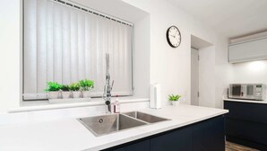 A modern kitchen with a stainless steel sink, a clock on the wall, and a window with blinds. - Host & Stay | Staffordshire Knot (Sheen)