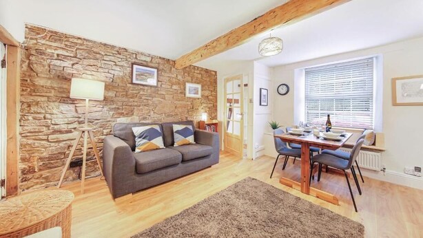 A cozy living room with a stone wall, a grey sofa, a dining table with chairs, and a large window with blinds. - Host & Stay | Batemill Cottage (Birch Vale)