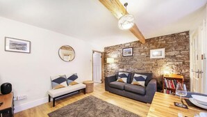 A cozy living room with a stone wall, a wooden ceiling, a grey sofa, a wooden coffee table, and a bookshelf. - Host & Stay | Batemill Cottage (Birch Vale)