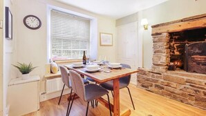 A dining area with a wooden table set for four, a stone fireplace, and a window with blinds. - Host & Stay | Batemill Cottage (Birch Vale)