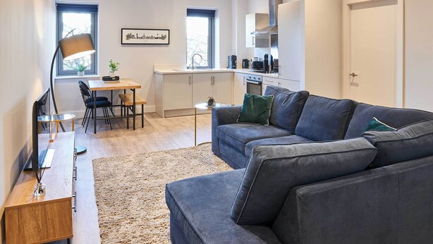 Una sala de estar moderna con un sofá gris, una mesa de centro de madera y un televisor.  Hay una zona de comedor con una mesa y sillas, y una cocina con armarios blancos y un ventanal.
