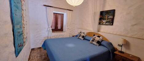 Un dormitorio con una cama grande, techo de madera, ventana con cortinas, un cuadro en la pared y una mesita de noche con lámpara.