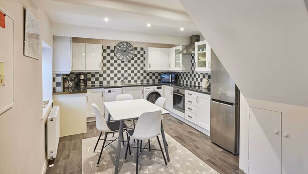 Eine moderne Küche mit weißen Schränken, einer schwarz-weißen Spritzschutzwand, einem Edelstahlkühlschrank und einem Essbereich mit weißem Tisch und Stühlen.