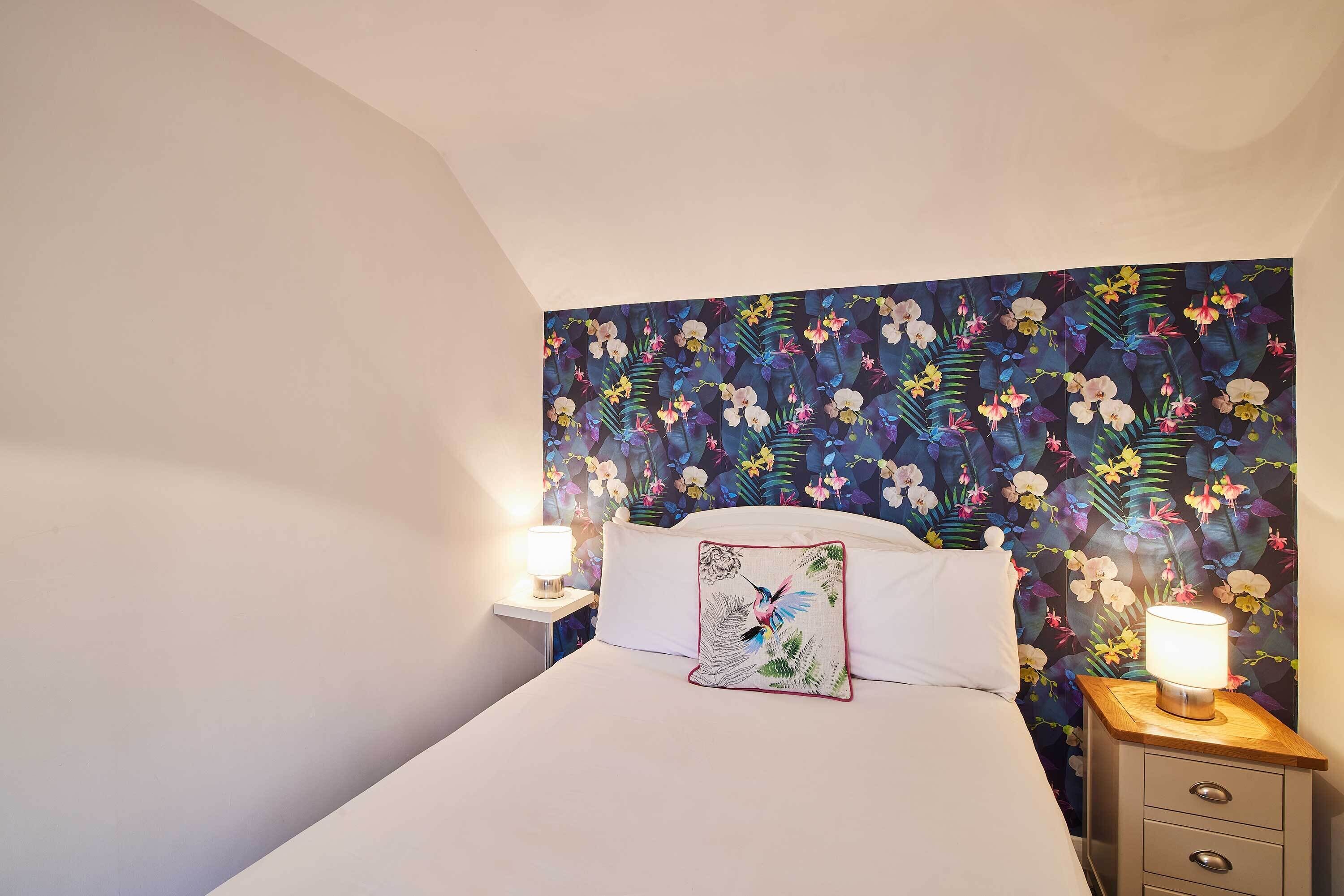 A bedroom with a floral wallpaper, a white bed, a decorative pillow, and two bedside lamps.