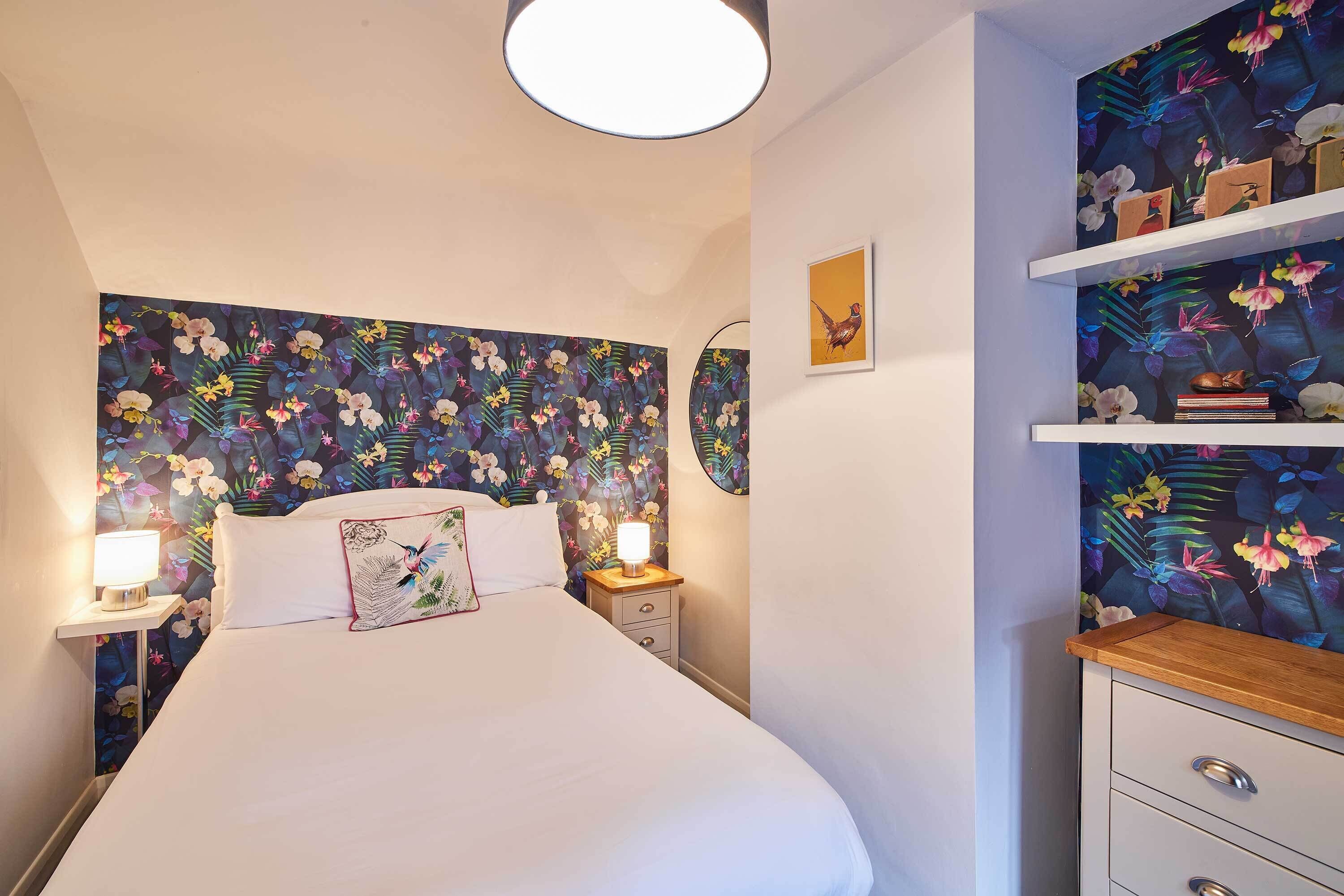 A bedroom with a floral wallpaper, a bed with white bedding, bedside lamps, a nightstand, and a wooden dresser with drawers.