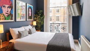 A neatly made bed with a white duvet and grey pillows, a bedside table with a lamp, and a large window with a view of buildings. - Host & Stay | The Falkner Quarters I and II (Merseyside)