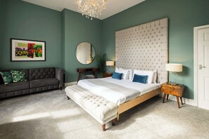 A bedroom with a large bed, a sofa, a round mirror, a chandelier, and a framed picture on the wall.