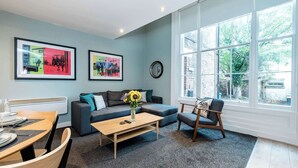 A modern living room with a sofa, a coffee table with a vase of flowers, a dining table set for two, and large windows offering a view of the outside. - Host & Stay | Georgian Quarters @ Canning I & II (Liverpool)