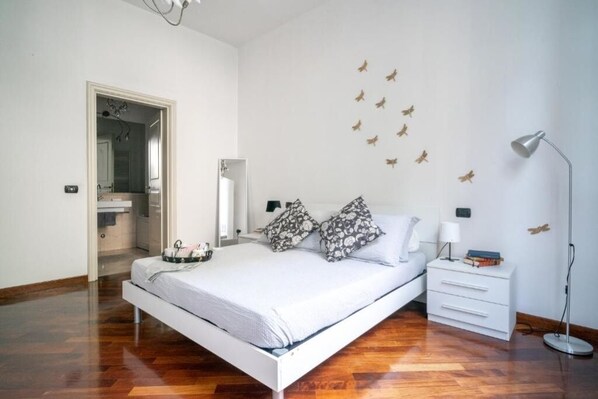 A bedroom with a white bed, a bedside table with books, a floor lamp, and a wall with bird decals.