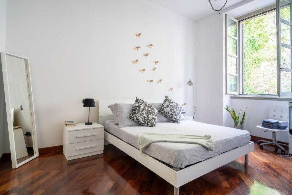 A bedroom with a bed, bedside table, lamp, and a window with a view of greenery.