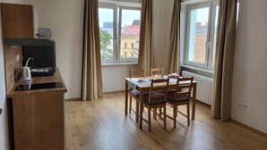 A room with a dining table, chairs, a kitchen area with a kettle and a stove, and large windows with curtains. - Apartmá Gabriela (Brno)