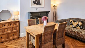 A dining area with a wooden table and wicker chairs, a sofa with patterned cushions, a mirror, a lamp, and a framed picture on the wall.