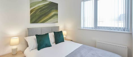A neatly arranged bedroom with a bed, bedside lamps, a wall art piece, and a window with blinds.