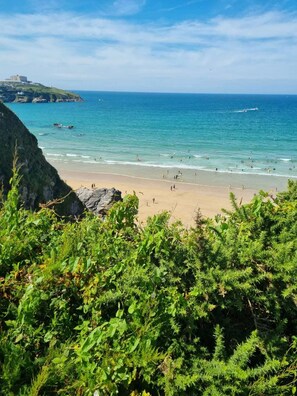 A beach with clear blue water, rocky cliffs, and lush greenery in the foreground. - Shamrock's Cove Caravan | Coastal Retreat | Family-friendly | Holiday Park (Newquay)