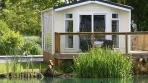 Ett litet hus med en träterrass som vetter mot en damm där det finns svanar.