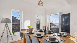 Un coin repas avec une table en bois dressée pour quatre personnes, un lampadaire sur trépied et une vue sur la mer grâce à de grandes fenêtres.