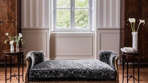 A room with a large window, a patterned sofa, two small tables with vases, and a wooden cabinet.