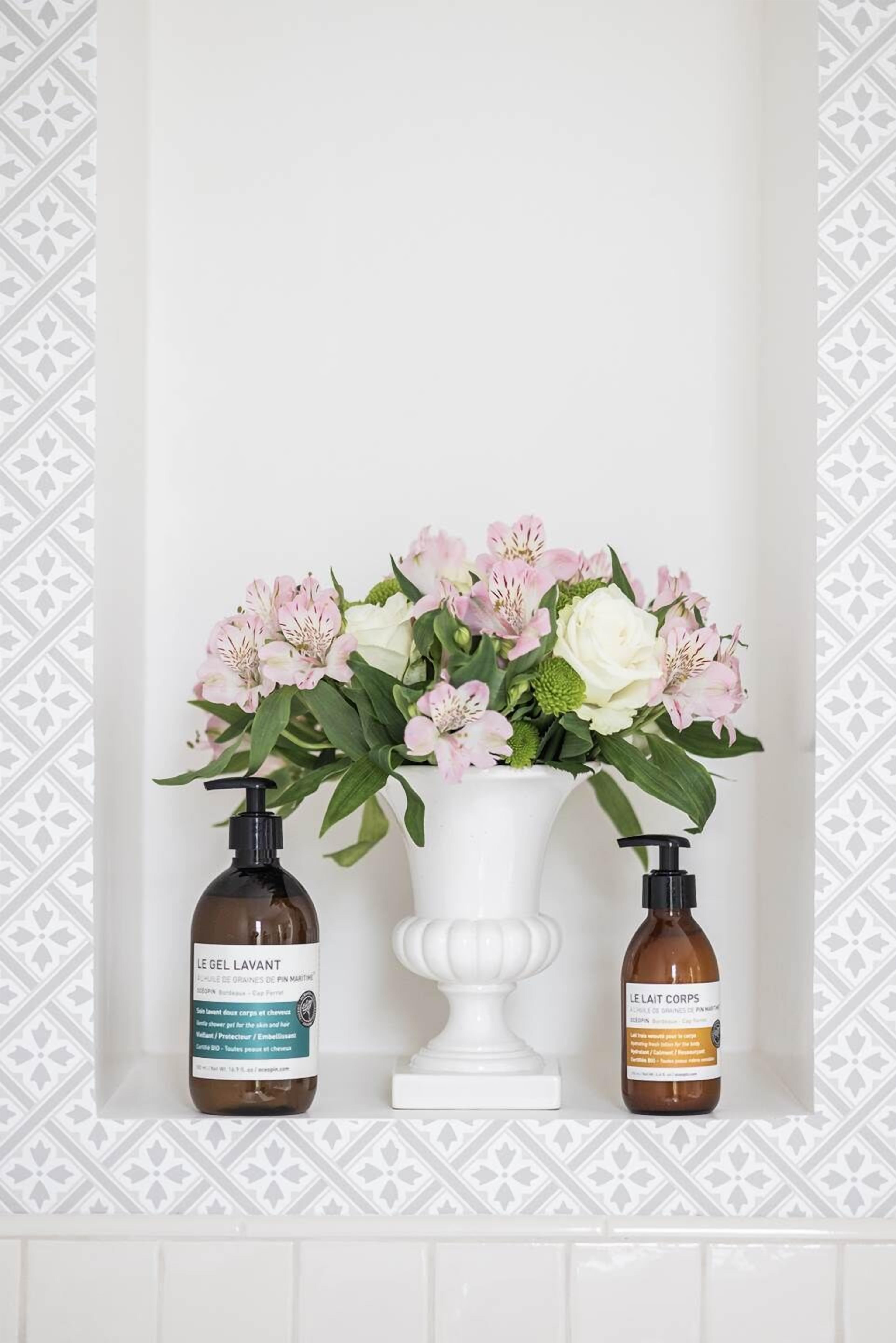 Une salle de bain avec un vase blanc contenant des fleurs roses et blanches, deux bouteilles de savon Le Lait Lavant et un lavabo sur pied blanc.