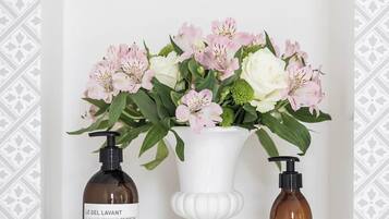 Une salle de bain avec un vase blanc contenant des fleurs roses et blanches, deux bouteilles de savon Le Lait Lavant et un lavabo sur pied blanc.