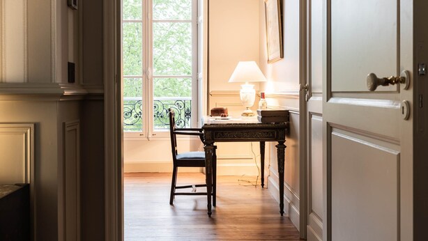 A hallway with a wooden floor, a chair, a table with a lamp, and a window with a view of greenery.