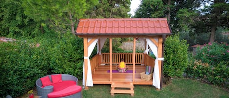 A wooden gazebo with a red roof, surrounded by lush greenery and a small seating area with red cushions.