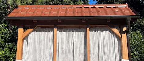 Un pabellón de madera con techo de tejas, cortinas blancas y un banco.