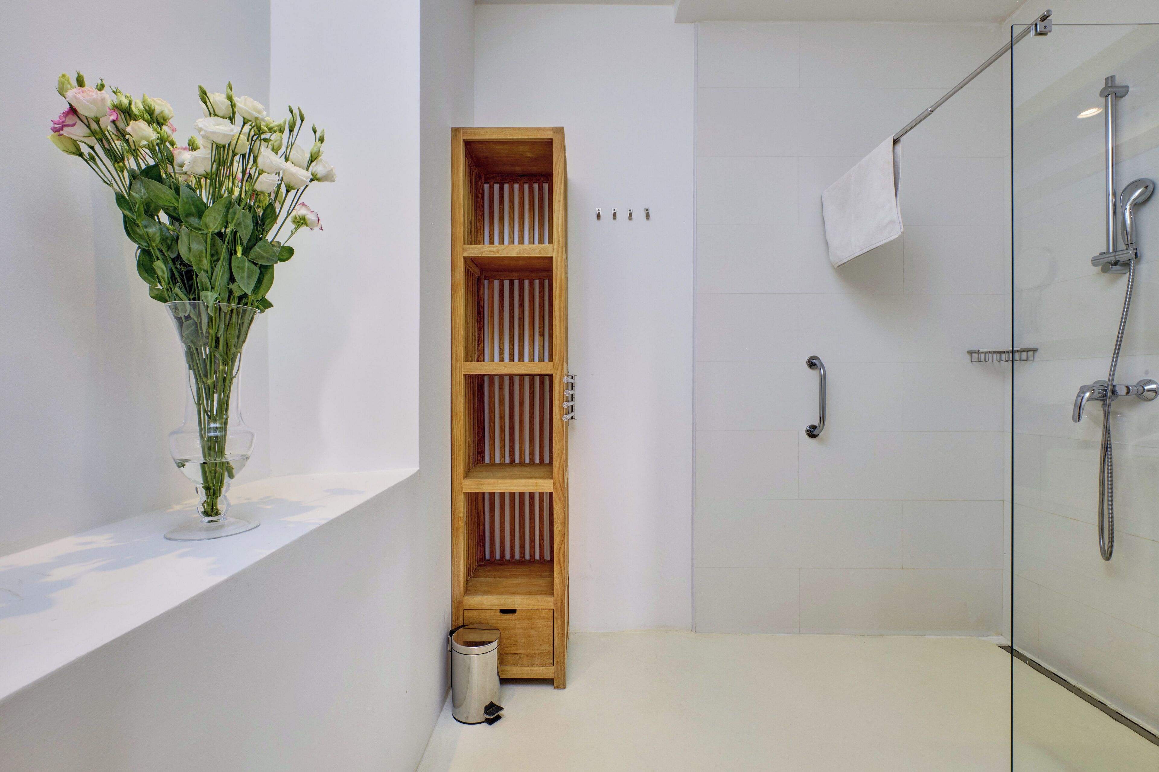 Une salle de bain moderne avec une douche à l’italienne en verre, une étagère en bois ornée d’un vase de fleurs et une petite poubelle.