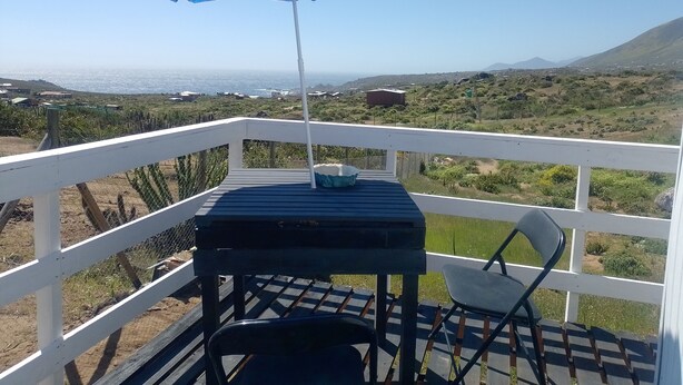 Un balcón con una sombrilla azul, una pequeña mesa y una silla, con vistas a un paisaje costero con casas y vegetación.