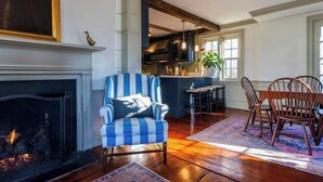 Un salon avec une cheminée, un fauteuil à rayures, une table à manger avec des chaises, et une cuisine en arrière-plan.