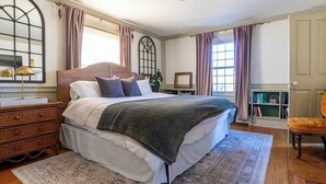 Une chambre à coucher avec un lit en bois, une table de chevet, une commode, une grande fenêtre avec des rideaux et une bibliothèque.
