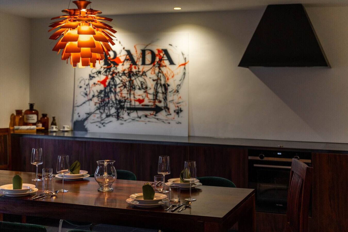 Ein Restaurantinnenraum mit einem für vier Personen gedeckten Esstisch, einer modernen Pendelleuchte und einer Wand mit einem großen abstrakten Gemälde.
