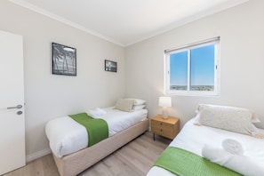 A hotel room with two beds, a window with a city view, a door, and a small bedside table with a lamp.