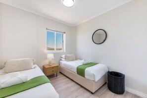 A bedroom with two beds, a window, a bedside table, a lamp, a mirror, and a trash bin. - Horizon Haven with stunning view by HMH (Cape Town)