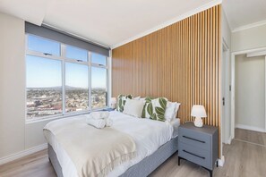 A bedroom with a large window, a bed with a white bedspread, a nightstand with a lamp, and a wooden slat wall. - Horizon Haven with stunning view by HMH (Cape Town)