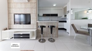 A modern kitchen with integrated space, a built-in TV, and a dining area with a table and chairs. - Comfortable apartment, complete infrastructure, Itacorubi, enjoy the beach and/or city! (Itacorubi Florianópolis)
