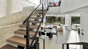 A modern interior with a staircase leading to a balcony, a dining area with a glass table, and a living room with a sofa and large windows.