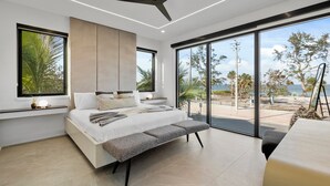 A modern bedroom with a large bed, a ceiling fan, and a view of the beach.
