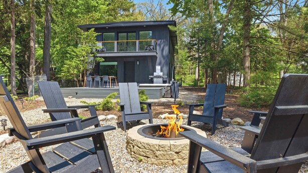 Un espacio moderno al aire libre con sillas Adirondack, un hogar para la leña y un edificio de dos pisos al fondo.