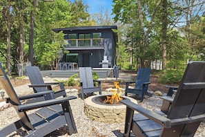 Un espacio moderno al aire libre con sillas Adirondack, un hogar para la leña y un edificio de dos pisos al fondo.