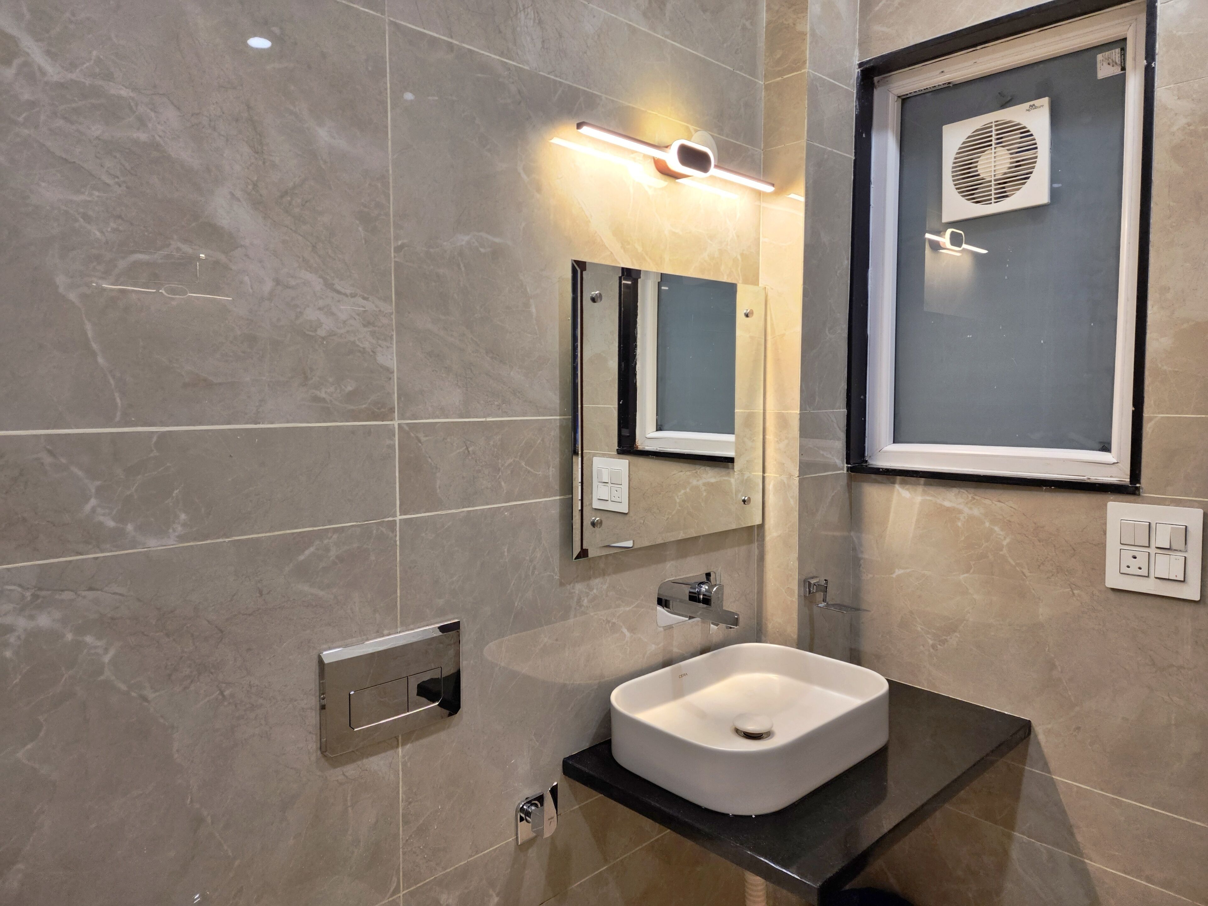 A modern bathroom with a white sink, a mirror, a wall-mounted light, and a window with a fan.