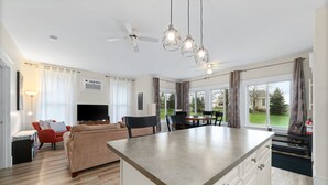 Una cucina moderna con isola centrale, zona pranzo con tavolo e sedie, soggiorno con divano e TV, e ampie finestre con tende.