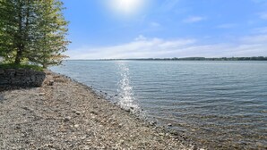 A pebbled beach with a stone wall and a tree, leading to calm water under a clear sky. - The Goldfinch - 36 Meadow View (Prince Edward)