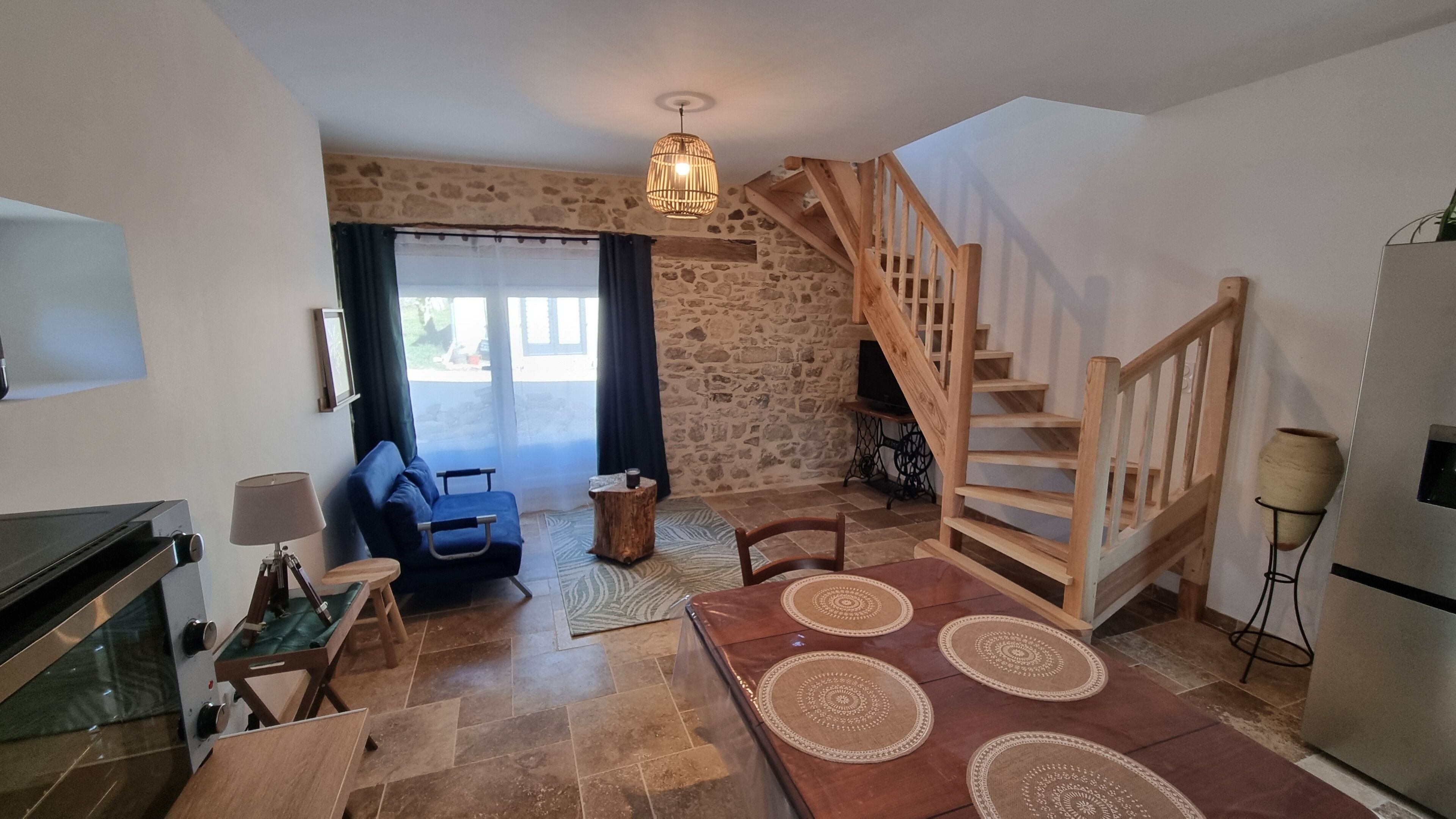 A cozy living room with a stone wall, a blue sofa, a wooden staircase, and a dining area with a table and chairs.