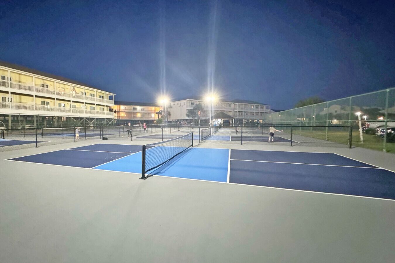 Ein Tennisplatz im Freien mit Netz, umgeben von einer Umzäunung und Gebäuden im Hintergrund.