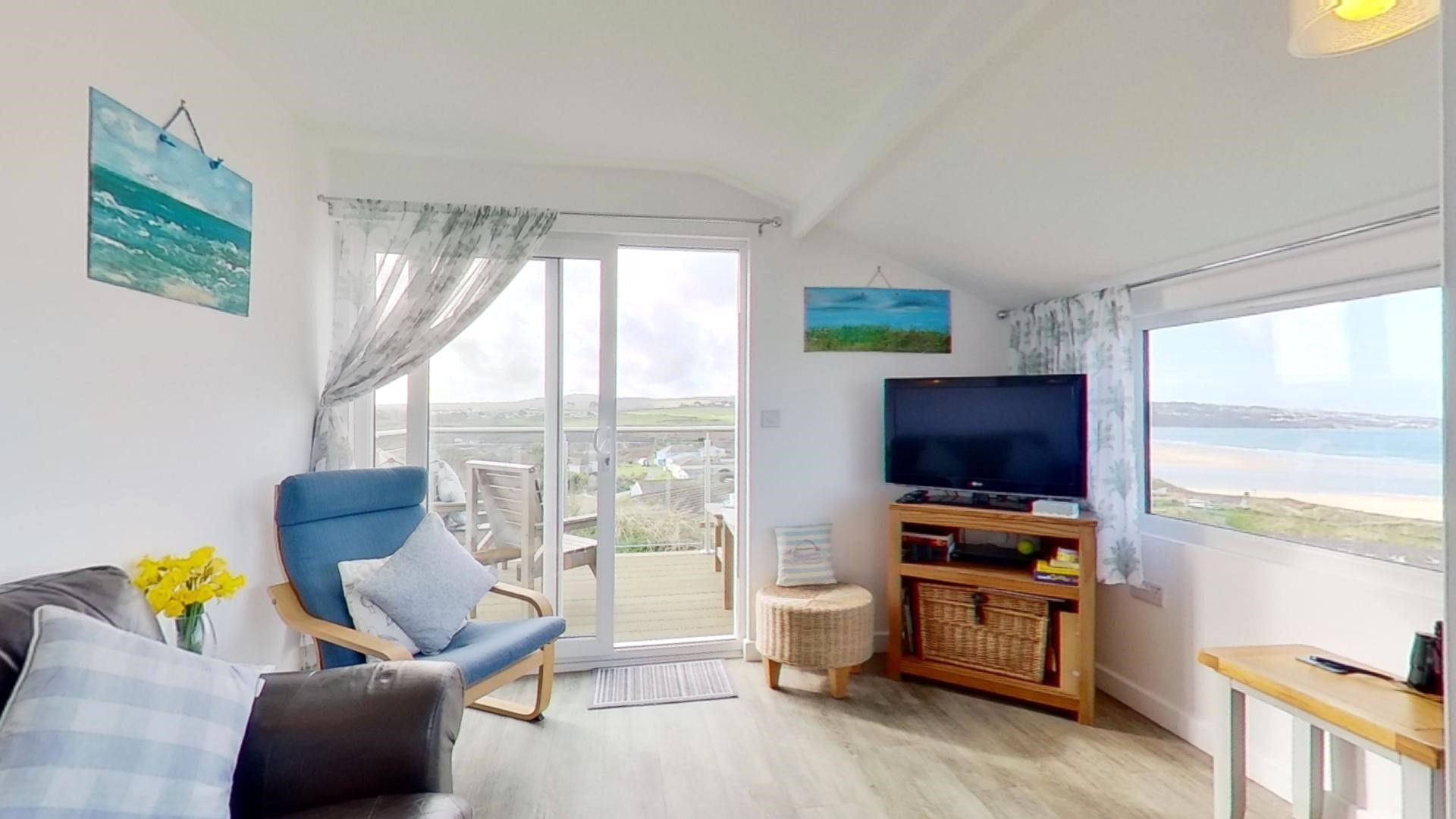 Un salon comprenant un canapé, un fauteuil, un téléviseur posé sur un meuble en bois, et une vue sur la mer grâce à de grandes fenêtres.